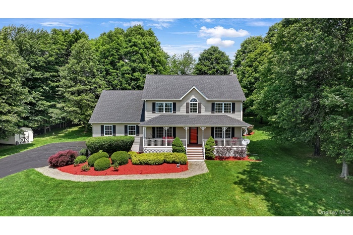 Colonial home with a porch, roof with shingles, a front lawn, asphalt driveway, and a storage unit and specimen plantings
