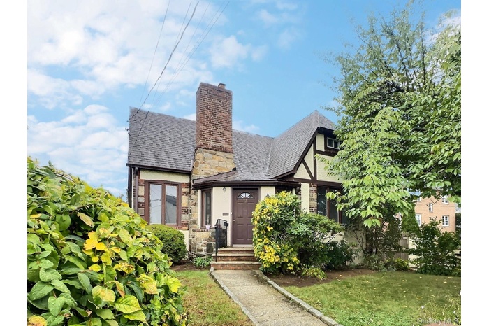 Tudor-style house with stone siding, a chimney, a front lawn, and a shingled roof