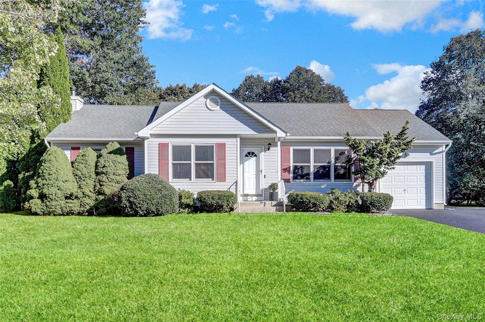 Ranch-style home featuring driveway, a front lawn, a shingled roof, an attached garage, and a chimney