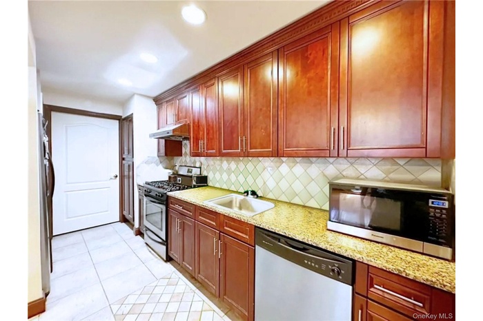 Kitchen with stainless steel appliances, light tile patterned flooring, backsplash, under cabinet range hood, and light stone counters