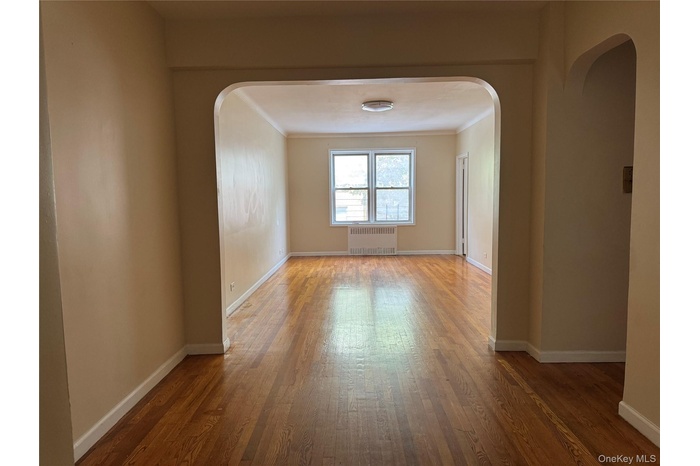 Empty room with arched walkways, wood finished floors, radiator heating unit, and ornamental molding