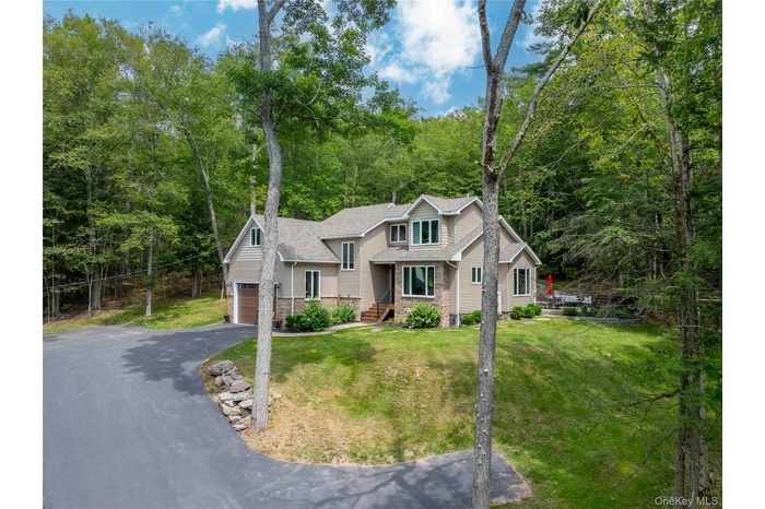 View of front of property featuring asphalt driveway, a front yard, a wooded view, and roof with shingles