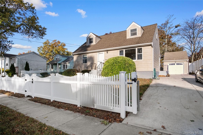Cape cod home featuring a fenced front yard, a shingled roof, a detached garage, an outdoor structure, and driveway