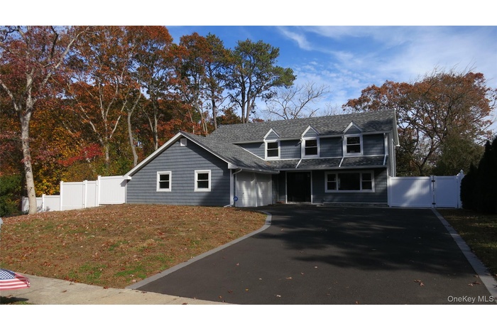 Cape cod home with a gate, asphalt driveway, a shingled roof, view of wooded area, and a garage