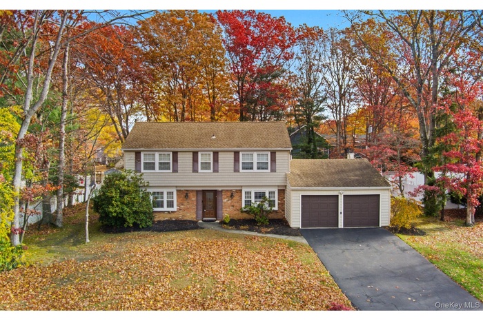 Colonial-style house featuring a shingled roof, brick siding, view of wooded area, asphalt driveway, and a front yard