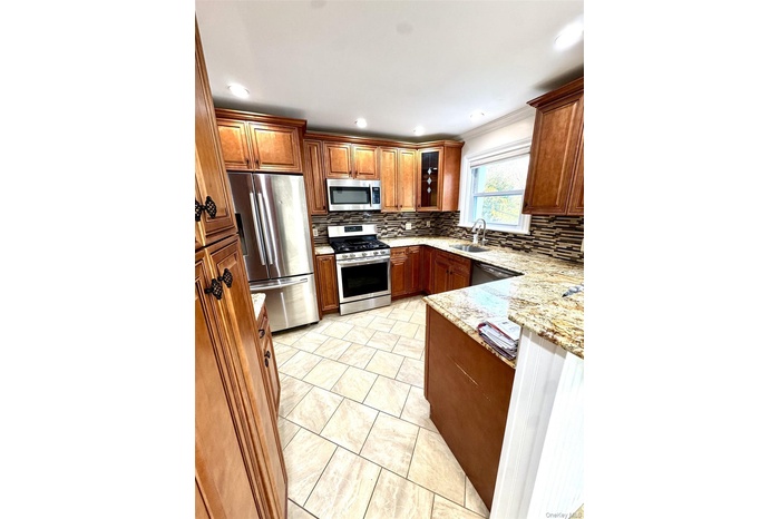 Kitchen featuring appliances with stainless steel finishes, brown cabinetry, light stone countertops, decorative backsplash, and glass insert cabinets