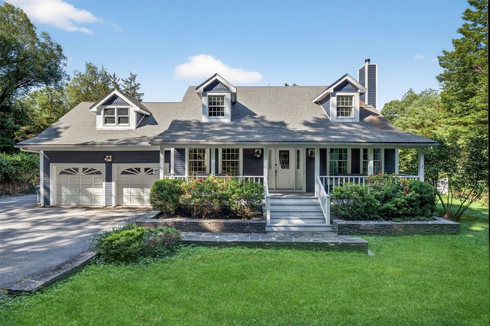 New england style home with a chimney, asphalt driveway, covered porch, a garage, and a front lawn