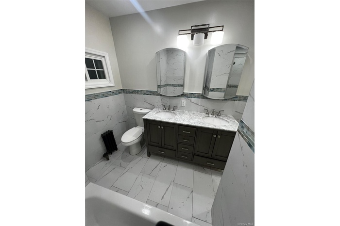 Bathroom featuring wainscoting, tile walls, double vanity, light marble finish flooring, and a tub