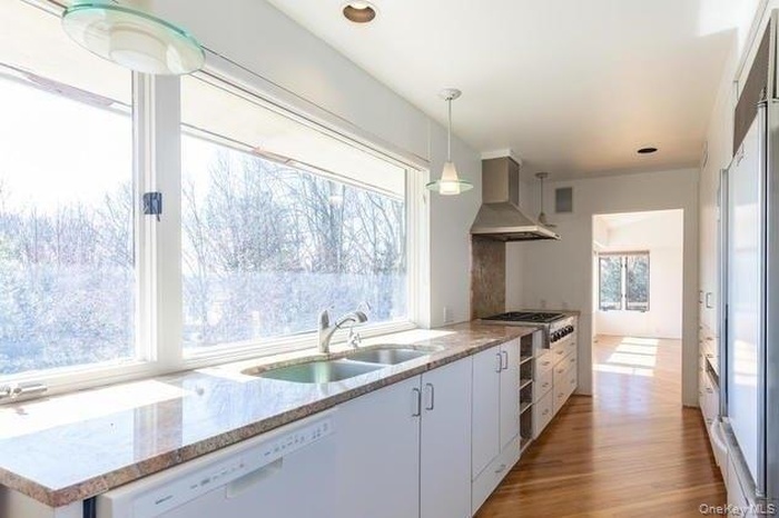 Kitchen with white cabinets, wall chimney exhaust hood, dishwasher, light wood-style floors, and hanging light fixtures