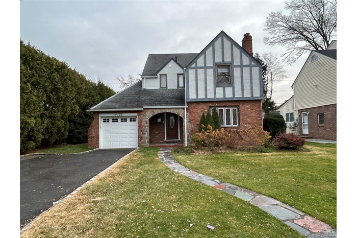 Tudor home with asphalt driveway, a front yard, brick siding, an attached garage, and a chimney