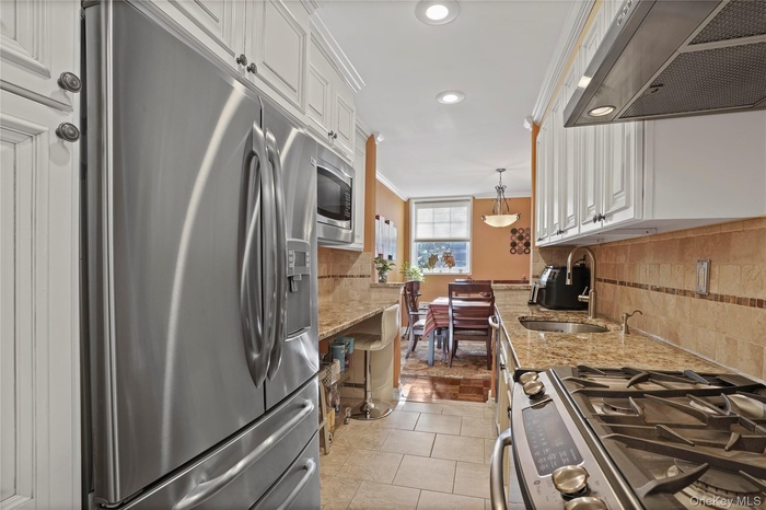 Kitchen featuring stainless steel appliances, light stone countertops, range hood, decorative light fixtures, and white cabinetry