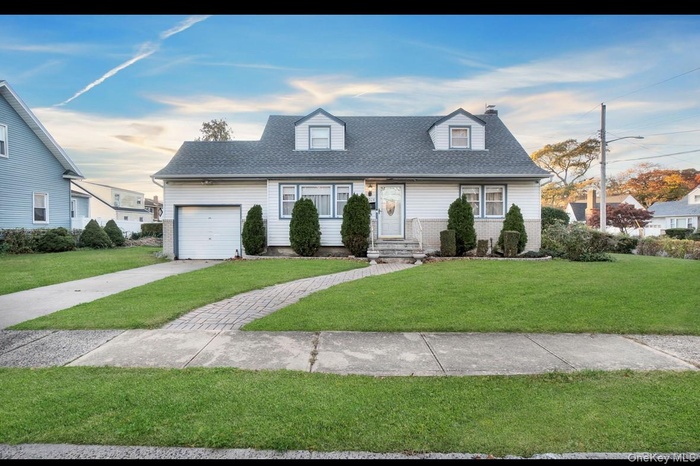 New england style home with a front yard, a shingled roof, a chimney, a garage, and driveway