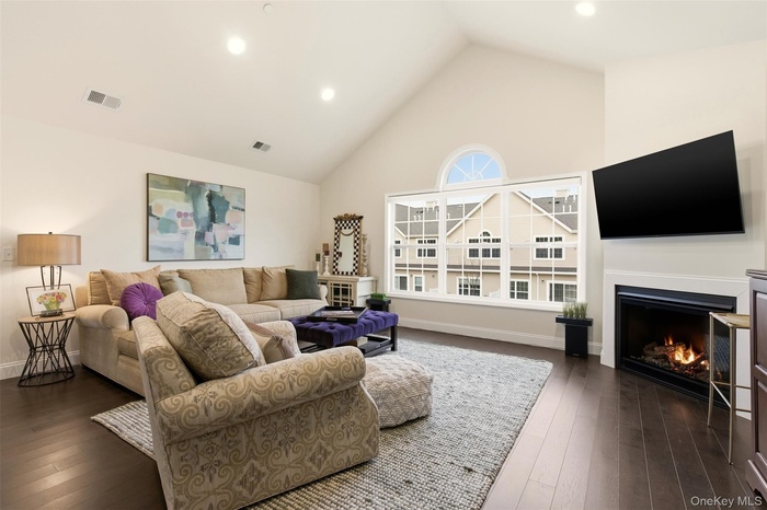 Living area with a warm lit fireplace, high vaulted ceiling, dark wood-style floors, and recessed lighting