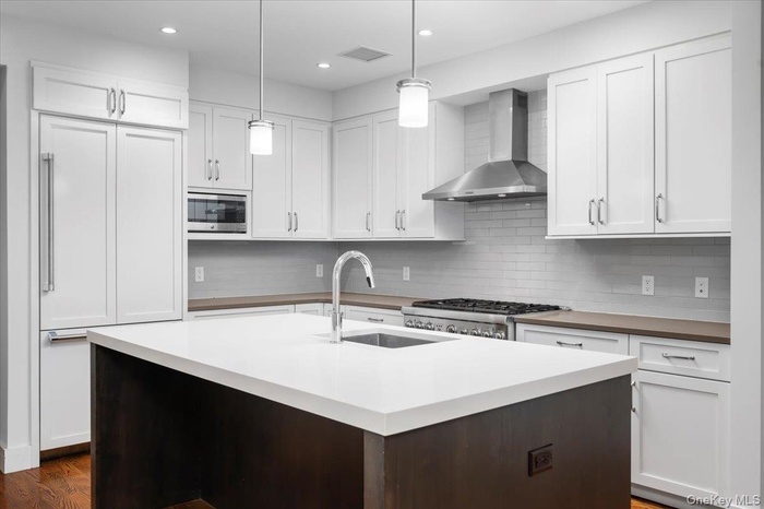 Kitchen with wall chimney range hood, white cabinetry, built in appliances, a center island with sink, and recessed lighting