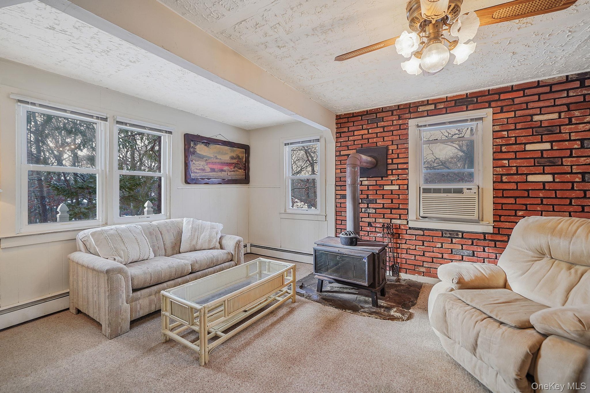 Carpeted living room with brick wall, a wood stove, a baseboard radiator, and ceiling fan
