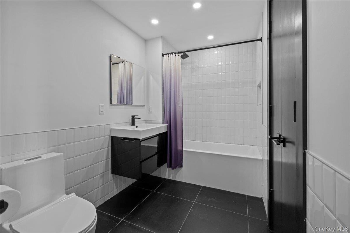 Bathroom featuring a wainscoted wall, tile walls, dark tile patterned floors, vanity, and shower / bathtub combination with curtain