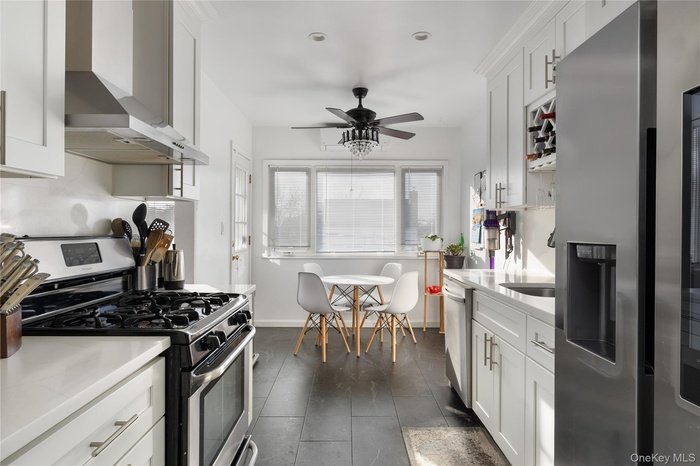 Kitchen featuring stainless steel appliances, wall chimney range hood, white cabinetry, and light stone countertops
