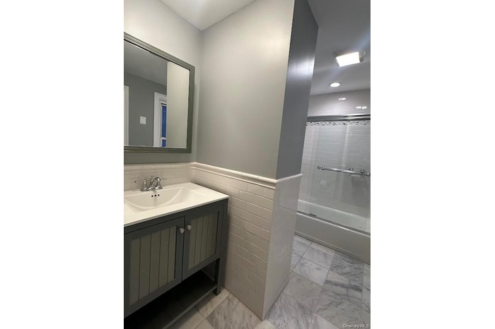 Bathroom featuring wainscoting, vanity, shower / bath combination with glass door, light marble finish flooring, and tile walls