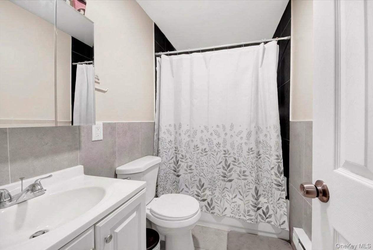 Bathroom with vanity, tile walls, a baseboard radiator, a wainscoted wall, and shower / bathtub combination with curtain