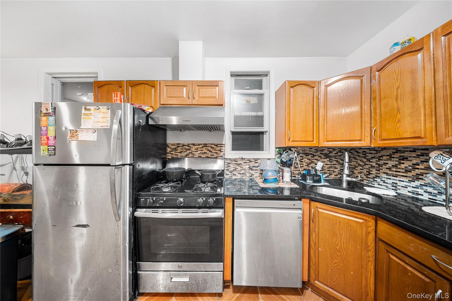 Kitchen with appliances with stainless steel finishes, brown cabinetry, under cabinet range hood, tasteful backsplash, and dark stone counters