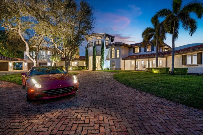 A striking twilight exterior showcasing the grand brick motor court, illuminated façade, and stately palm-lined approach. Sophisticated and commanding