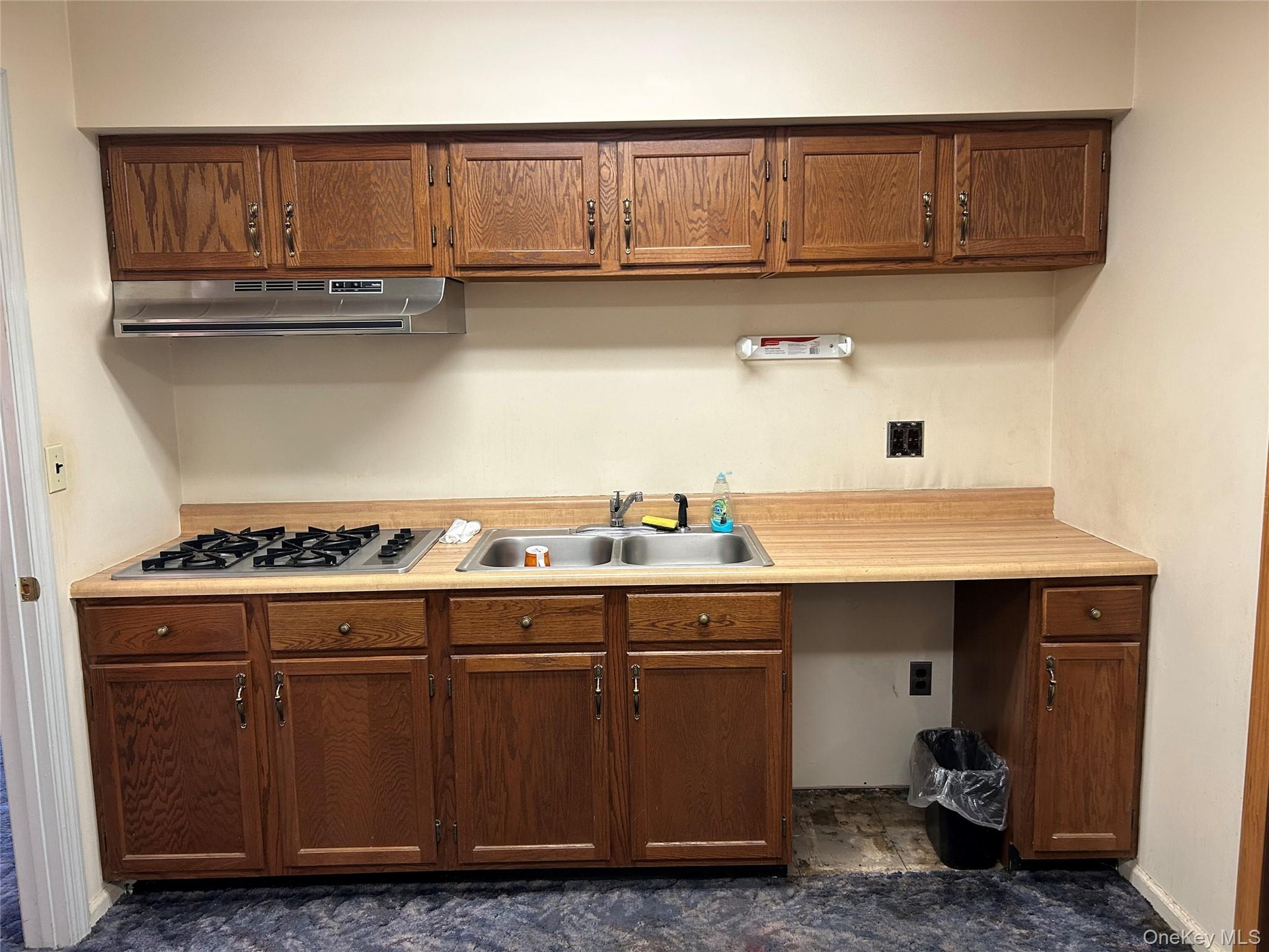 Kitchen featuring light countertops, under cabinet range hood, stainless steel gas stovetop, brown cabinetry, and dark stone finish flooring