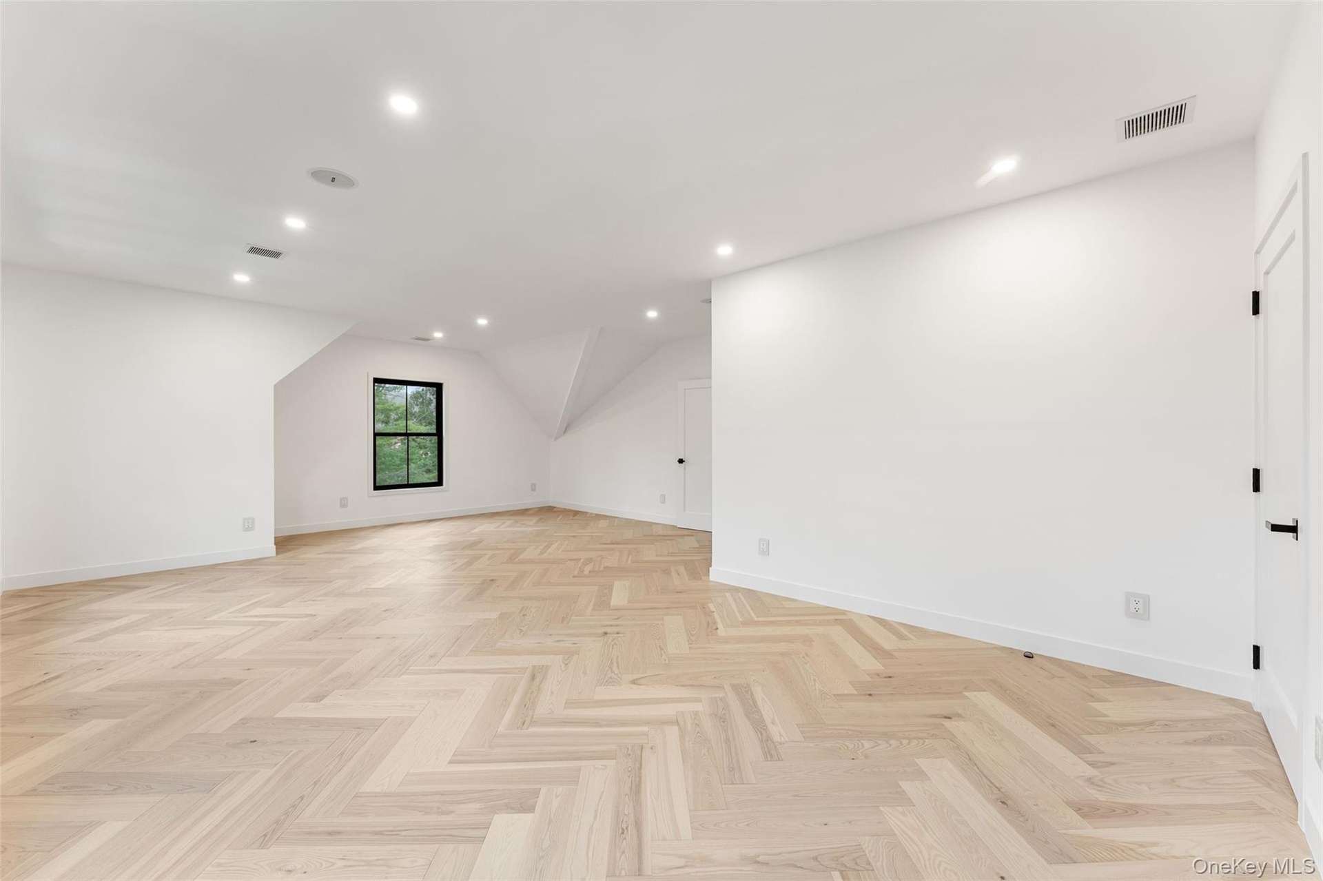 This versatile bedroom features light-toned hardwood flooring in a herringbone pattern, recessed lighting, and a window providing natural light