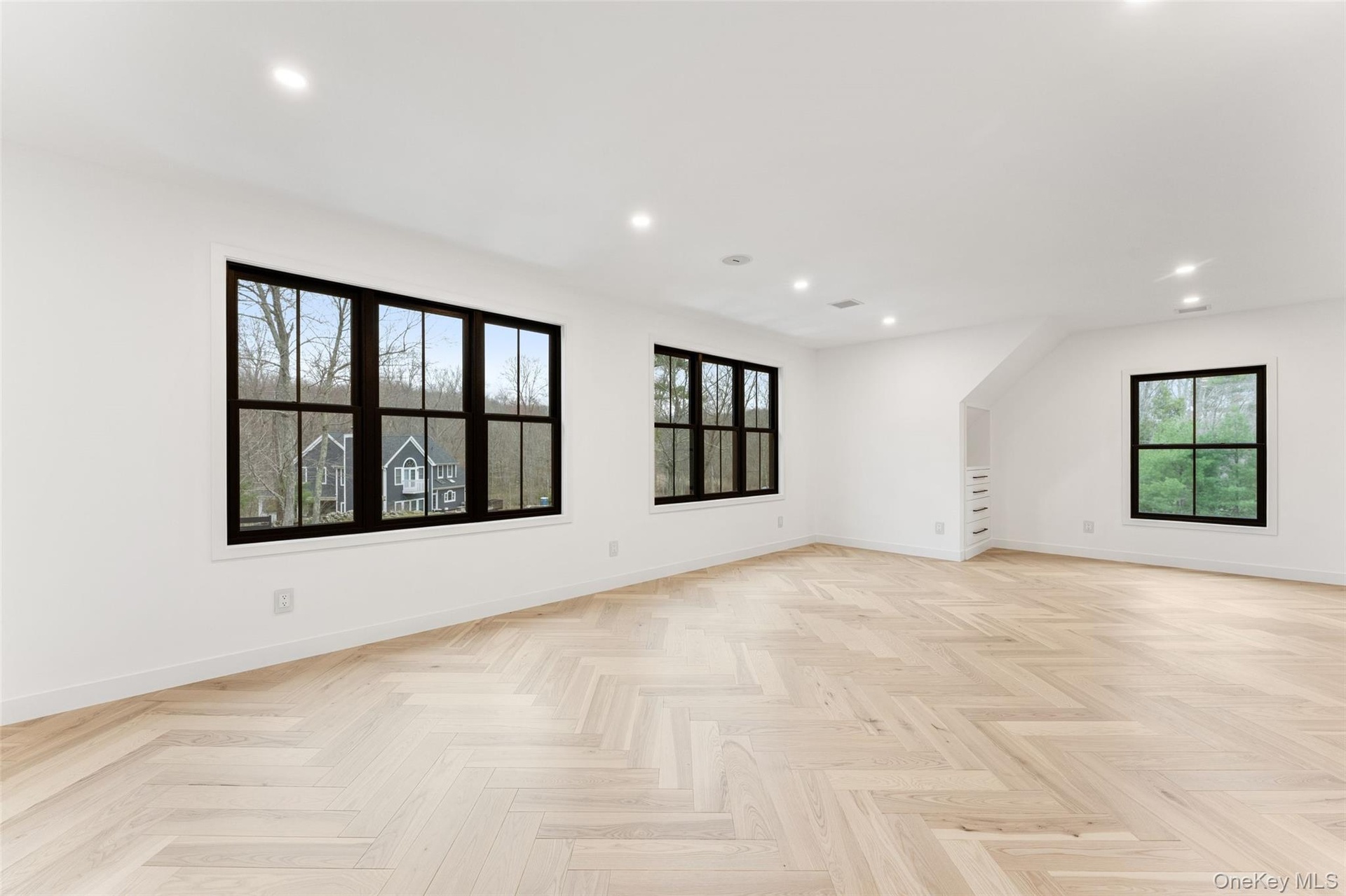 Expansive bedroom featuring light hardwood floors laid in a herringbone pattern, recessed lighting, and multiple windows with dark frames