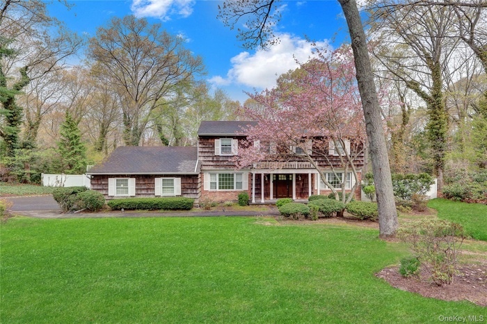 Colonial-style exterior featuring a wood shingle facade, a covered front porch with white columns, and a mature flowering tree