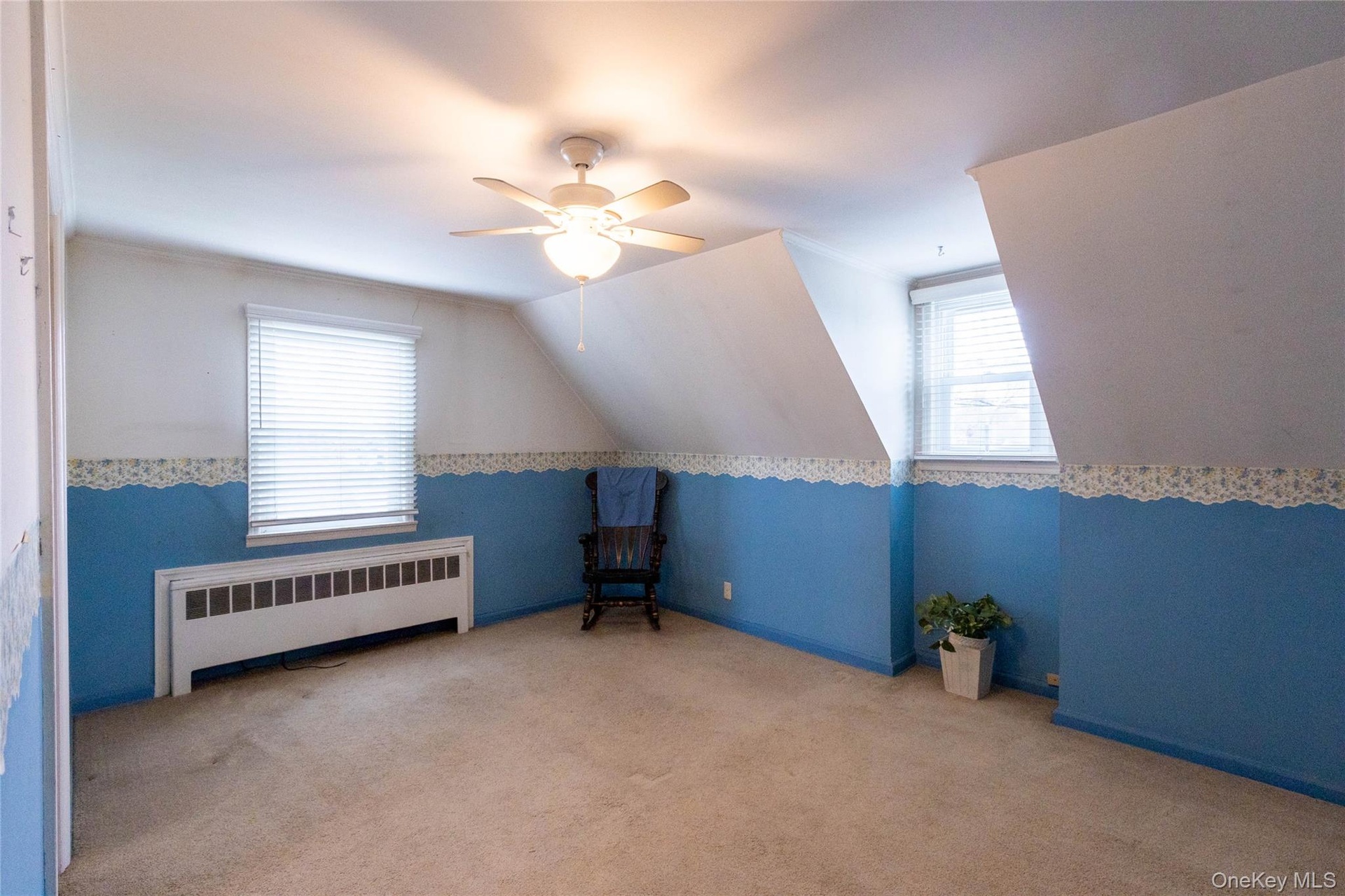 Carpeted room featuring a ceiling fan with integrated lighting, two windows with horizontal blinds, an integrated heating unit, and sloped ceiling det