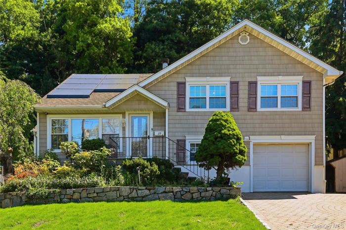 Shingle-sided residence featuring solar panels, a stone retaining wall, and a patterned paver driveway