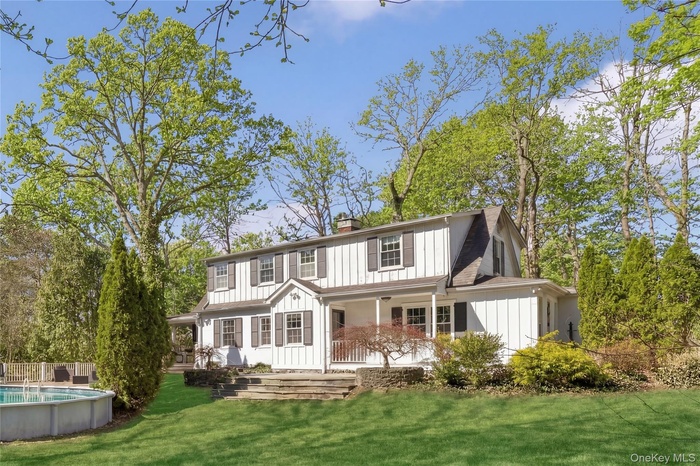 White facade residence featuring a covered porch, dark window shutters, dormer windows, a manicured lawn, and a round above-ground pool