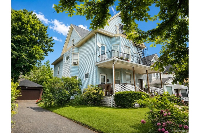 Light blue siding exterior featuring multiple balconies with black iron railings, a covered porch with white lattice detailing, a paved driveway, and 
