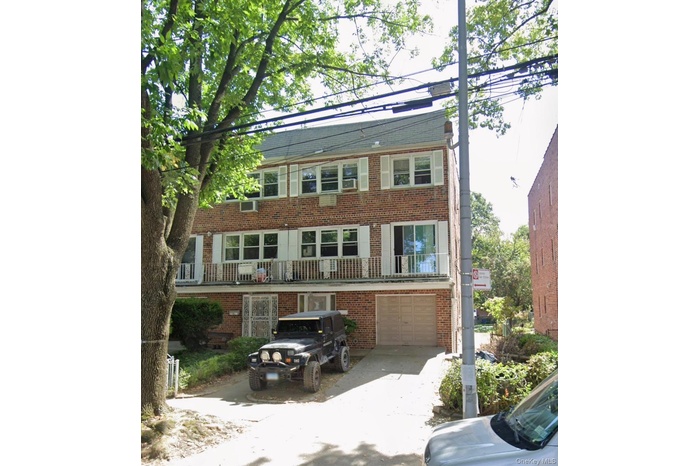 Brick-clad multi-story building featuring multiple balconies with white railings, white louvered shutters, an integrated garage, and a concrete drivew