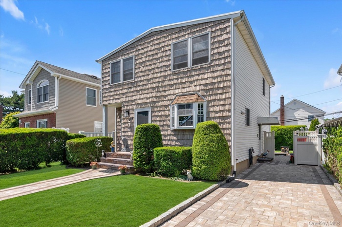 Two-story residence featuring vinyl siding exterior, bay window, front entry with brick steps, paver driveway, and manicured lawn with mature hedges,