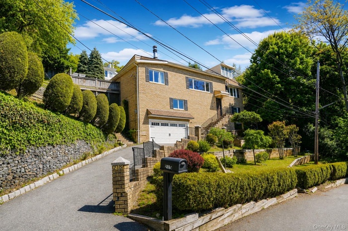 Brick façade residence featuring an integrated garage, a terraced front yard, established hedges, and a paved driveway with stone retaining walls