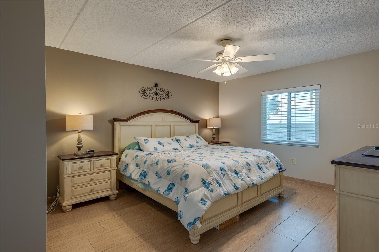 This spacious bedroom offers warm neutral tones, a ceiling fan for added comfort, and a large window that brings in natural light. The room’s layout a