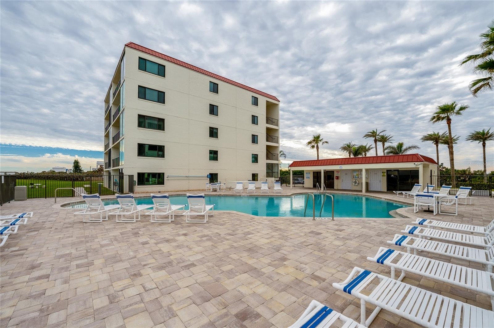 The resort-style pool includes a spacious deck, poolside lounge chairs, and a nearby clubhouse building, all set against a backdrop of palm trees and 