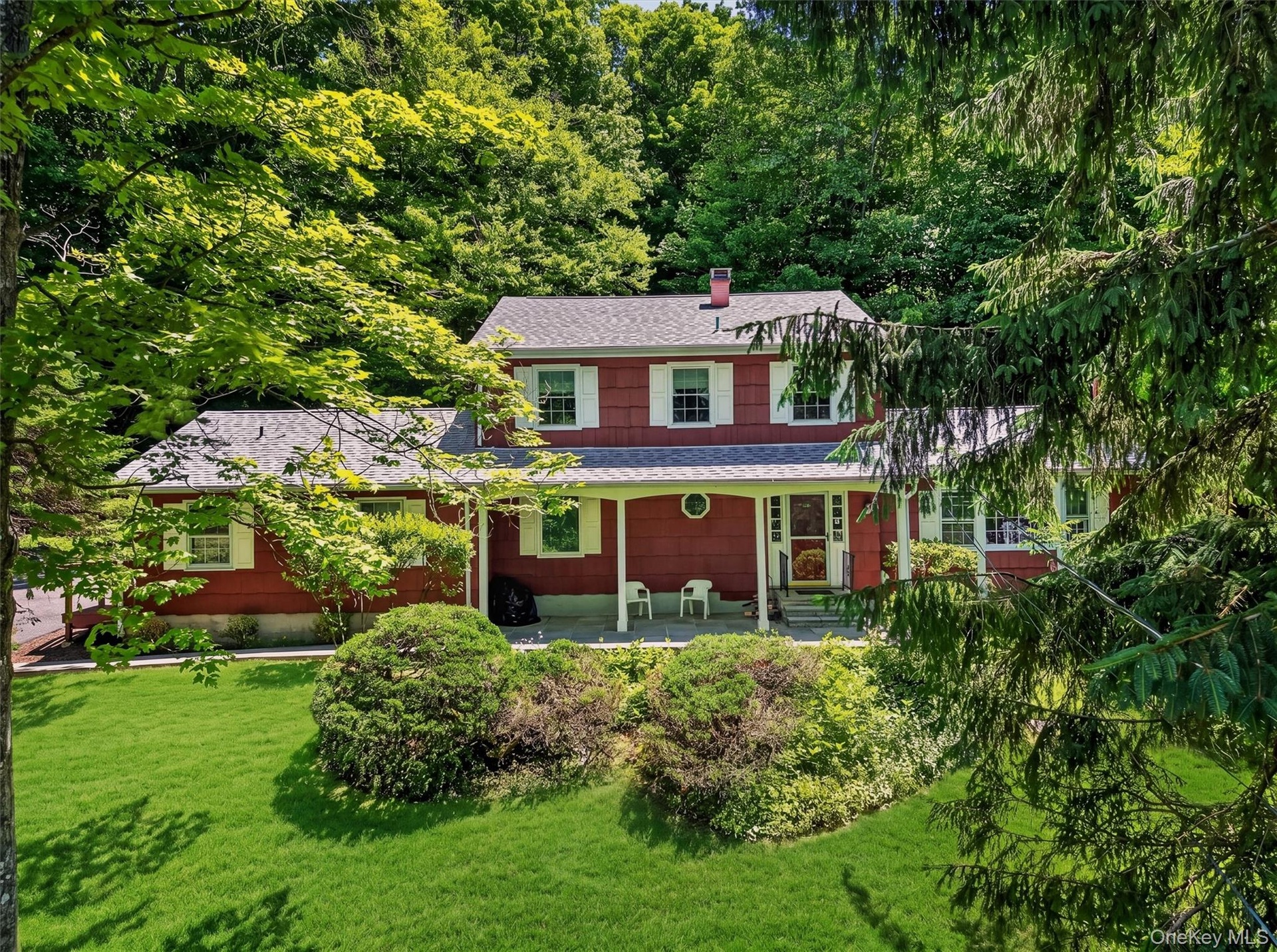 Residence featuring covered front porch, white window shutters, a grey shingled roof, and a verdant lawn with mature trees and shrubs
