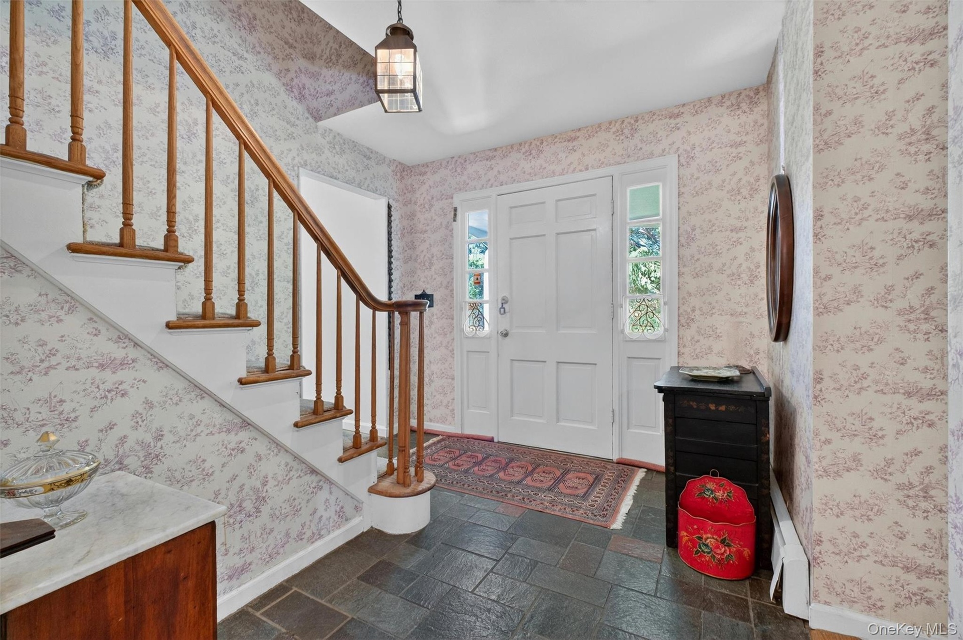 Foyer featuring raised panel front door with side light windows, dark slate tile flooring, a curved wood handrail, and patterned wallpaper