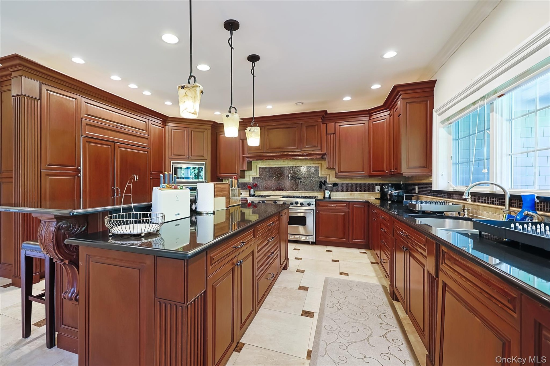Spacious kitchen featuring rich wood cabinetry, dark countertops, a large center island with pendant lighting, integrated appliances, and light-toned 