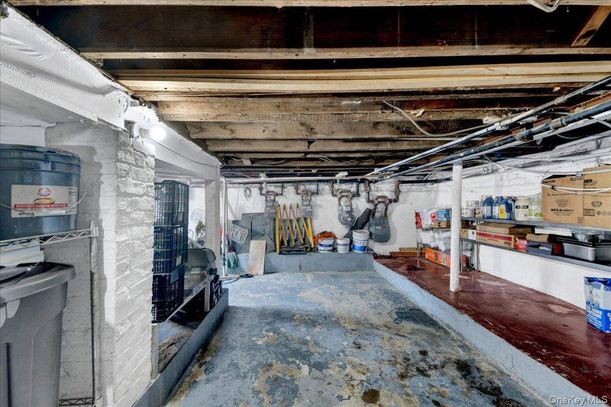 Storage space with exposed ceiling joists and support beams, featuring a painted brick wall, concrete flooring, and visible utility lines