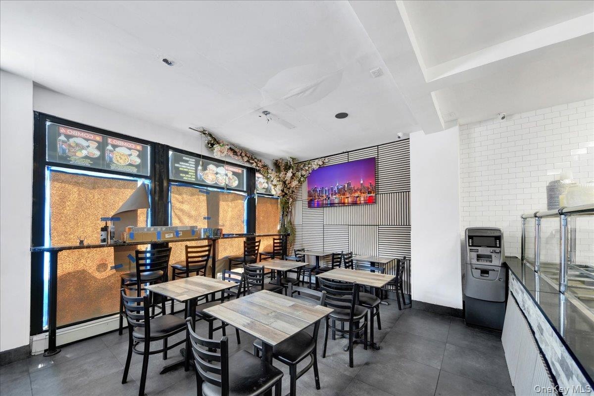 Commercial dining area featuring multiple tables and chairs, white subway tile wall, large display screen, and a service counter with glass enclosure