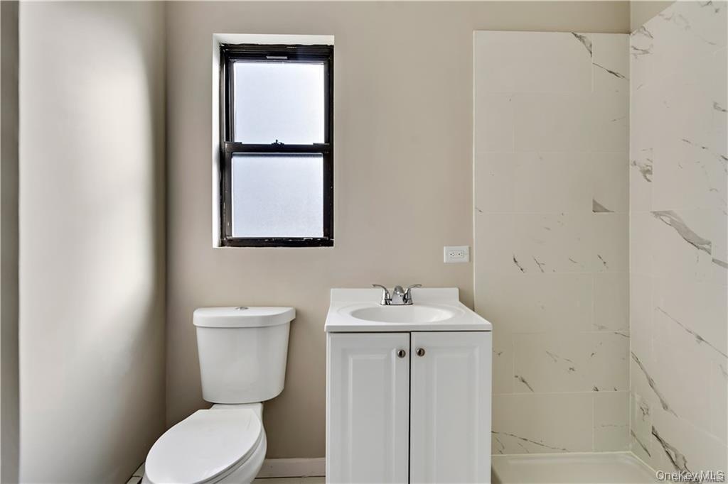 Bathroom featuring a single-pane window, white toilet, vanity with integrated sink, chrome faucet, and large-format porcelain tile shower surround