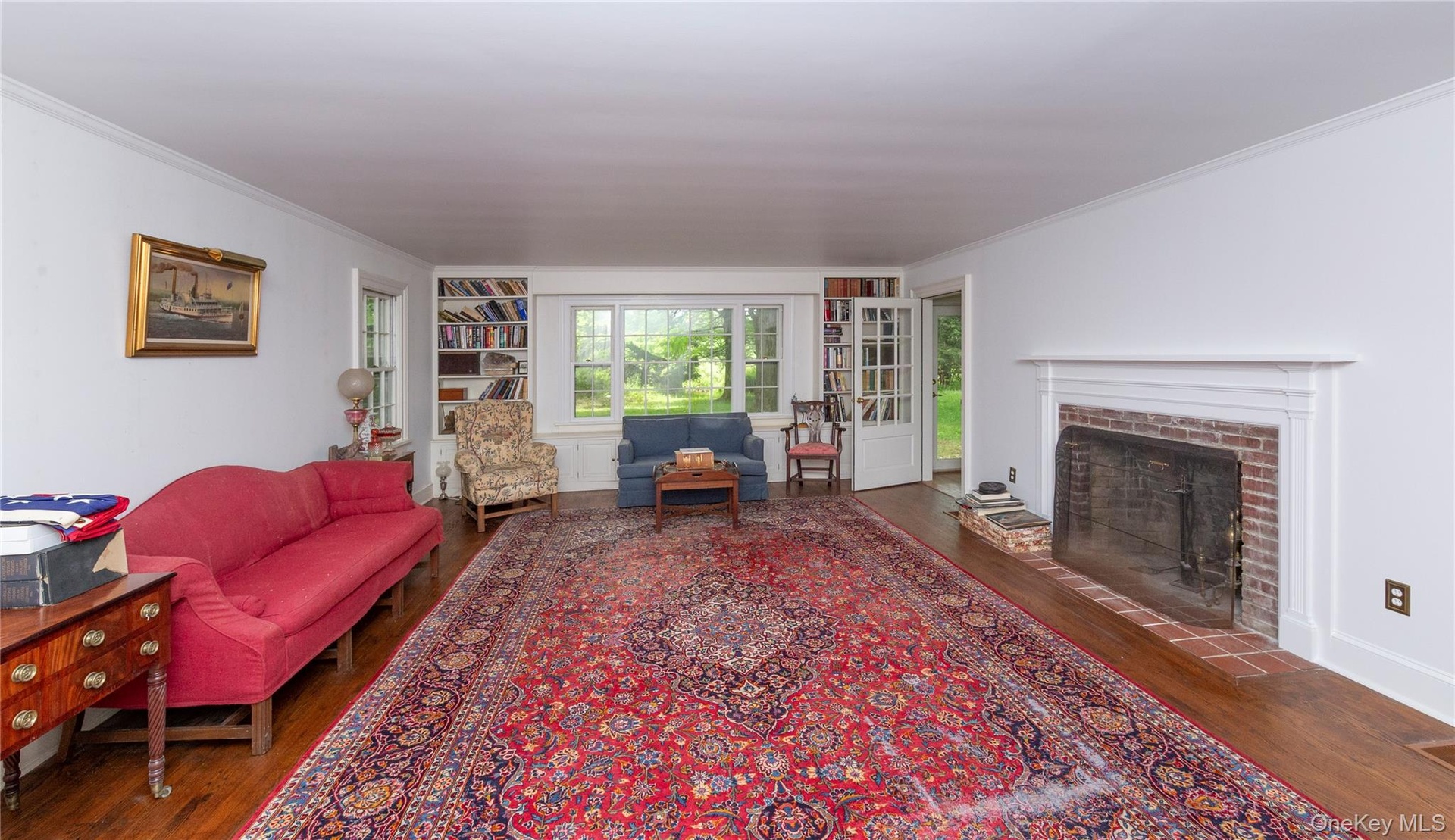 Spacious interior room featuring wood-finish flooring, a brick fireplace with a white mantel, and built-in shelving flanking a large window