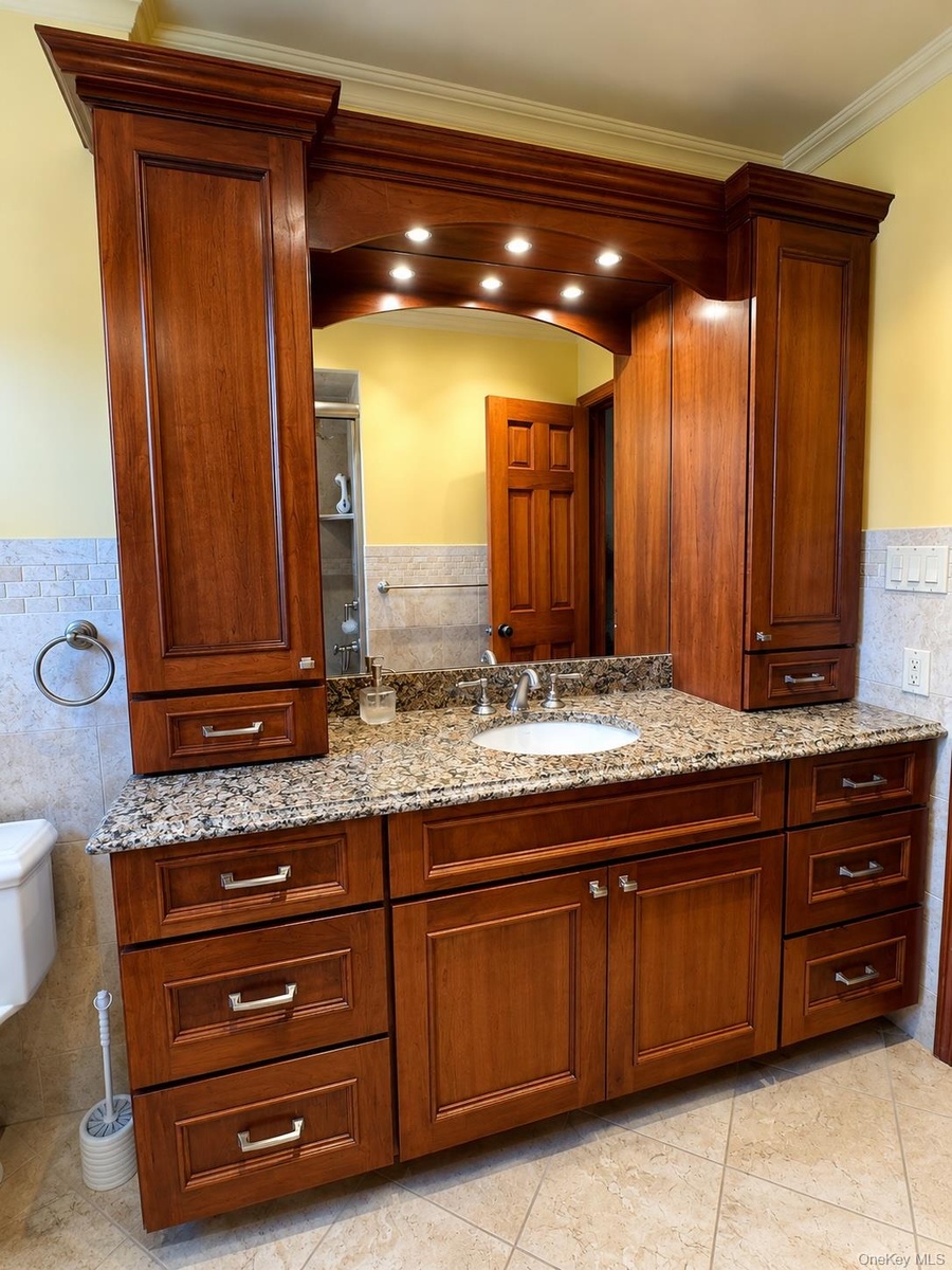 Custom vanity featuring cherry-toned cabinetry, granite countertop, undermount sink, and integrated overhead lighting