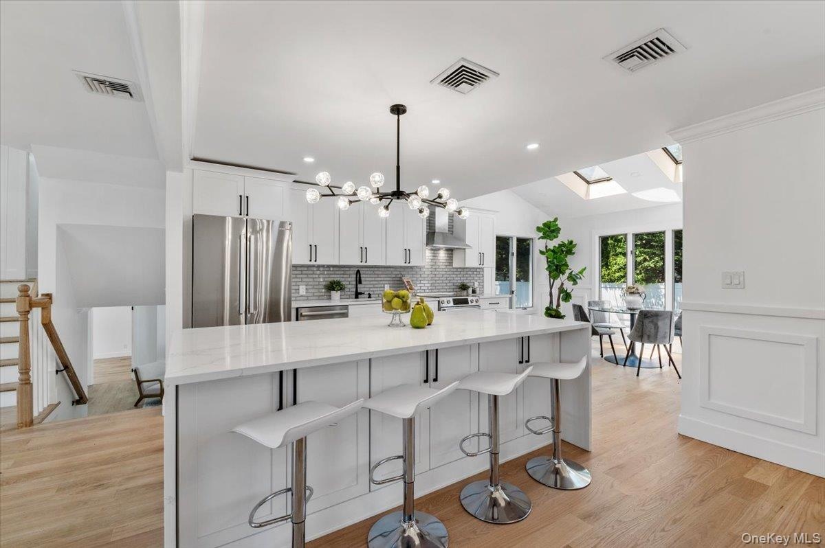 Kitchen featuring freestanding refrigerator, wall chimney range hood, decorative backsplash, white cabinetry, and a skylight