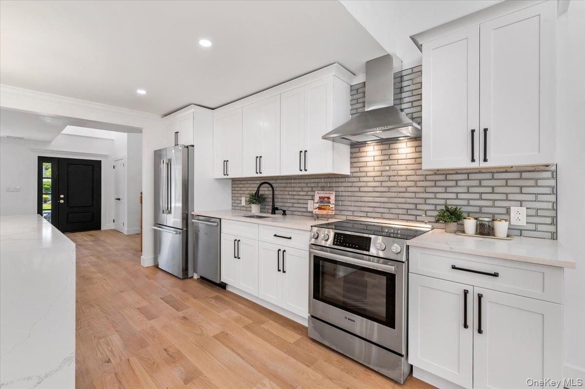Kitchen with wall chimney range hood, stainless steel appliances, white cabinetry, backsplash, and recessed lighting