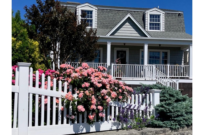 Gambrel roofline property featuring a full-width front porch with white columns, light grey shingle siding, and two dormer windows. Perennial rose bus