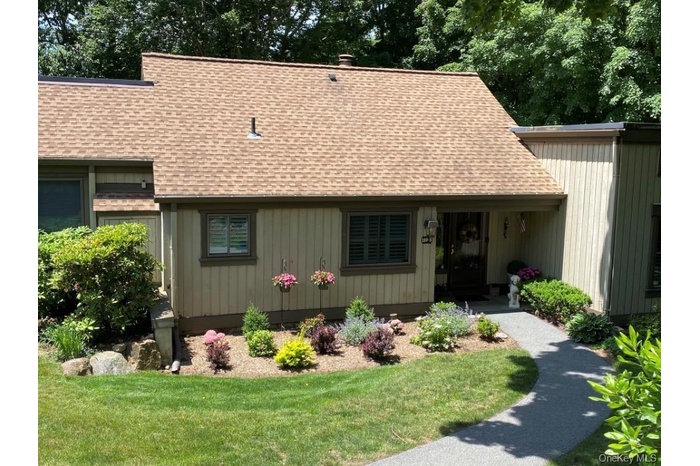 Exterior featuring a composite shingle roof, vertical siding, a paved walkway, established landscaping, and a covered entryway